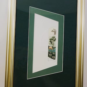 May include: A framed print with a green mount and a gold frame. The print features a landscape scene with a small animal in the foreground.