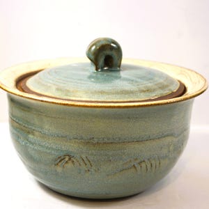 May include: A handmade ceramic bowl with a lid. The bowl is a muted teal colour with brown and cream accents around the rim and lid. The lid has a small, rounded handle. The bowl has a textured surface with etched designs.