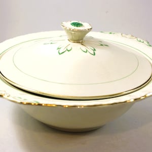 May include: Cream-coloured serving bowl with lid. The bowl has a scalloped edge with gold trim. The lid features a green floral design and a small, decorative handle. The bowl is designed for serving food.