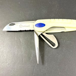 May include: A folding multi-tool knife with a cream-colored handle, a serrated stainless steel blade, a marlinspike, and a shackle key. The blade features the text Wichard and a logo. A blue oval emblem with a logo is on the handle.