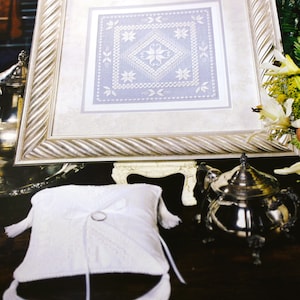 May include: A white satin ring pillow with a silver ring on top. The pillow is sitting on a table with a silver teapot and a framed piece of embroidery.