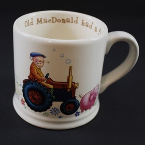 May include: White ceramic mug with a cartoon illustration of a farmer driving a tractor with a pig and sheep in the background. The mug has the text "Old MacDonald had a f..." printed on the side.