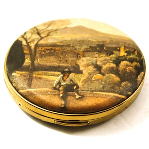 May include: A round, gold-toned compact with a landscape scene on the lid. The image depicts a man sitting on a wall, with a town and mountain in the background. The compact is closed and has a shiny finish.