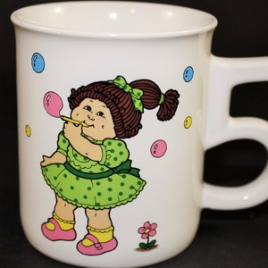 May include: White ceramic mug featuring a cartoon girl blowing a bubble. The girl wears a green polka-dot dress and has brown hair. The mug has a unique handle and is decorated with colorful bubbles and a flower.