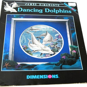 May include: Cross stitch pattern for a round design featuring four dolphins swimming in a blue ocean with coral reefs. The pattern is titled "Dancing Dolphins" and is by James Himsworth for Dimensions.
