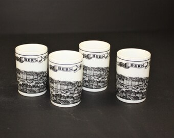 May include: Four white ceramic containers with lids. Each container features a black and white cityscape design and the word "BERN" on a banner. The containers are cylindrical and sit on a dark surface.