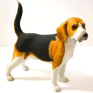 May include: A Beagle dog figurine with a tri-colour coat. The dog has a black back, with tan and white markings on its body, head, and legs. The tail is raised, and the dog appears to be standing, with a detailed finish.
