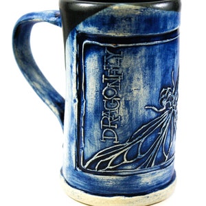 May include: A blue ceramic mug with a dragonfly design and the word "Dragonfly" embossed on the side. The mug has a black rim and a white base.