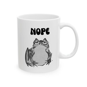 May include: White ceramic mug with a grumpy frog illustration in grey tones. The word "NOPE" is printed above the frog in bold, black letters. The mug has a curved handle for easy gripping.