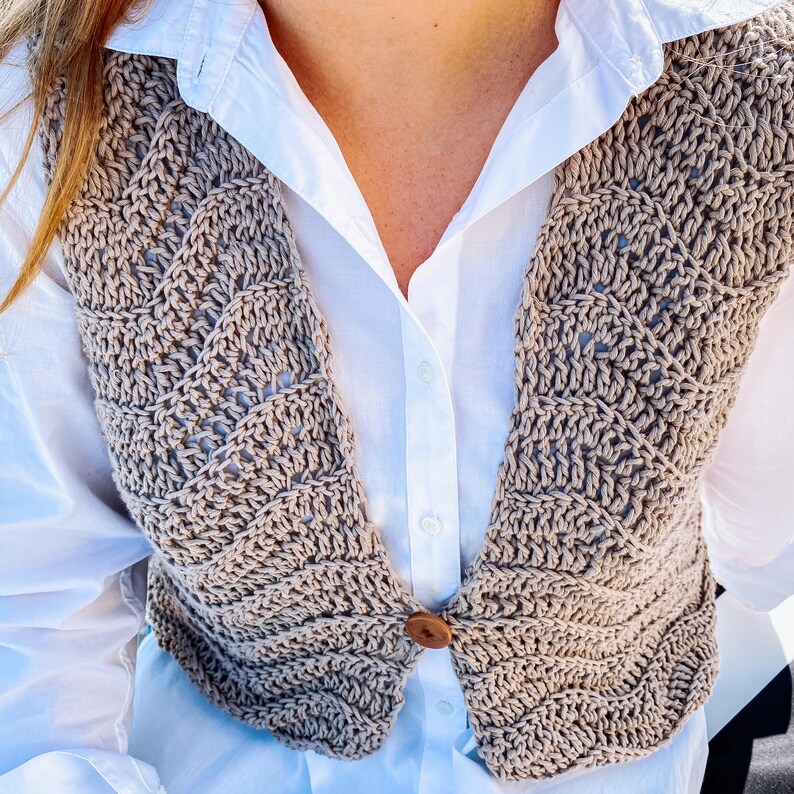 Pu&ograve; includere: Un gilet all'uncinetto color beige caratterizzato da un motivo a punti ondulati e testurizzati con una chiusura a bottone marrone al centro. Il gilet &egrave; indossato sopra una camicia bianca con colletto, maniche lunghe e abbottonatura.