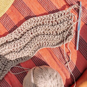 Pu&ograve; includere: Un progetto all'uncinetto beige in corso con un motivo a punti ondulati, accompagnato da un gomitolo di filato abbinato e un uncinetto in metallo argentato. Il filato &egrave; lavorato a righe, mostrando la consistenza dei punti e il filo libero.