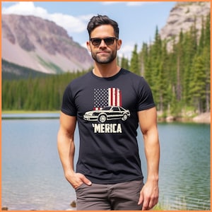 May include: A black t-shirt with a vintage American flag design and a black muscle car with the text 'MERICA' underneath. The model is wearing the shirt and standing in front of a lake and mountains.
