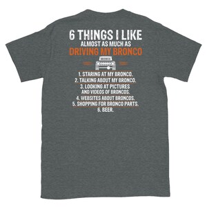 May include: A dark heather grey t-shirt with a white graphic that says "6 Things I Like Almost As Much As Driving My Bronco". The graphic also includes a black and white illustration of a Ford Bronco.