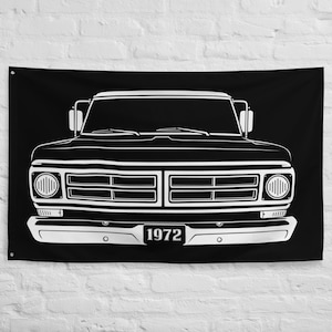 May include: Black wall flag featuring a white outline of a vintage 1972 pickup truck. The truck's front view is detailed, including headlights, grille, and the license plate with the year. The flag is rectangular and hangs against a textured white wall.