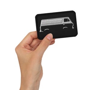 May include: A black embroidered patch with a white outline of a van. The patch is held in a hand.