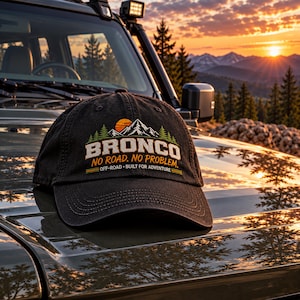 May include: A black baseball cap featuring a graphic of a mountain range, pine trees, and a sun. The text reads BRONCO, NO ROAD. NO PROBLEM., and OFF-ROAD BUILT FOR ADVENTURE. The cap has a curved brim and stitched detailing on the front panel.