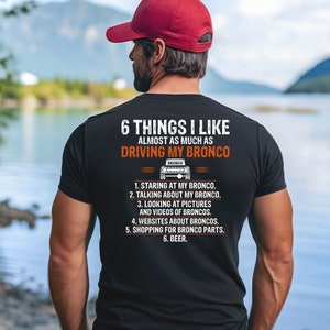 May include: A black t-shirt with a white graphic that says "6 Things I Like Almost As Much As Driving My Bronco". The graphic also includes a small image of a Ford Bronco.