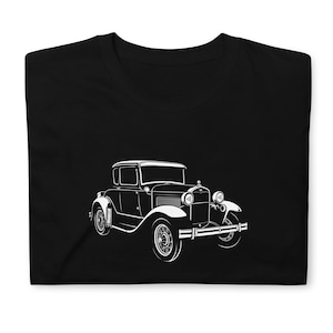 May include: Black t-shirt featuring a white illustration of a vintage car. The classic car design includes details like headlights, a radiator grille, and rounded fenders. The shirt is folded, showcasing the graphic prominently.