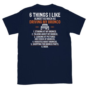 May include: A navy blue t-shirt with a white graphic of a Ford Bronco and the text "6 THINGS I LIKE ALMOST AS MUCH AS DRIVING MY BRONCO". The text below the graphic lists six things the wearer likes almost as much as driving their Bronco: "1. STARING AT MY BRONCO. 2. TALKING ABOUT MY BRONCO. 3. LOOKING AT PICTURES AND VIDEOS OF BRONCOS. 4. WEBSITES ABOUT BRONCOS. 5. SHOPPING FOR BRONCO PARTS. 6. BEER."