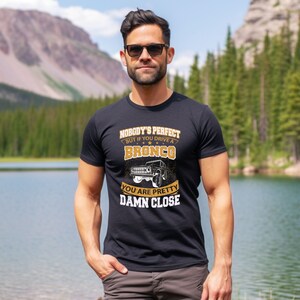 Peut inclure: Un t-shirt noir avec un motif doré qui dit "Nobody's perfect but if you drive a Bronco you are pretty damn close". Le motif comprend également une illustration en noir et blanc d'un Ford Bronco.