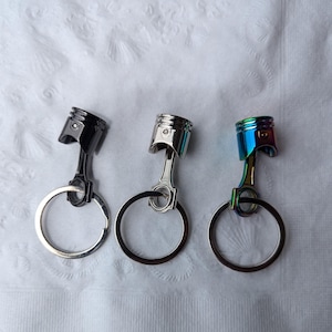 May include: Three metal keychains shaped like a piston. The keychains are silver, black, and iridescent blue.