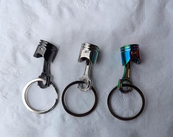 May include: Three metal keychains shaped like a piston. The keychains are silver, black, and iridescent blue.