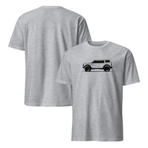 Peut inclure: Deux t-shirts gris clair. Le t-shirt avant présente un dessin au trait noir d'un véhicule tout-terrain. Le véhicule est une vue de profil avec de grandes roues et une forme carrée. Le dos du t-shirt est uni.