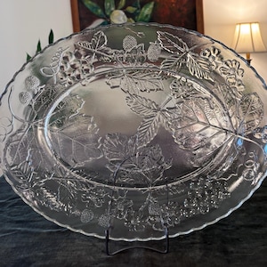 Op de afbeelding: Een ovale serveerschaal van helder glas met een verhoogd, reliëfpatroon van aardbeien, bladeren en wijnranken. De schaal heeft een geschulpte rand en een glad, verzonken midden, met een gedetailleerd botanisch ontwerp in het glas geperst.