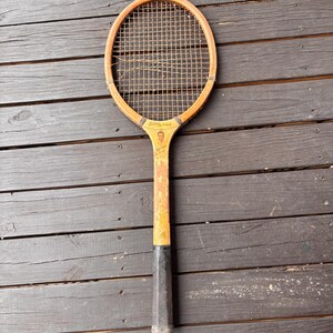 May include: A vintage wooden tennis racket with a black grip and a yellow painted surface. The racket has a stringed head and a faded image of a person on the handle.