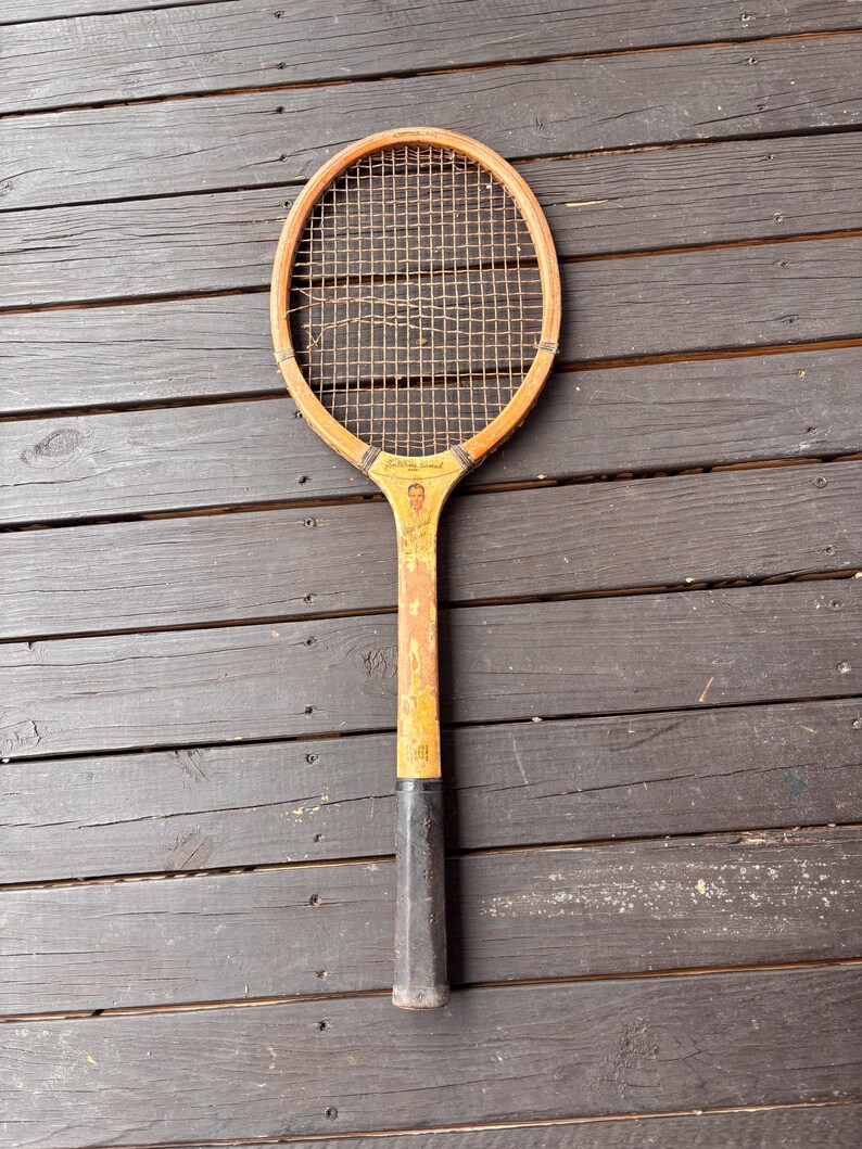 May include: A vintage wooden tennis racket with a black grip and a yellow painted surface. The racket has a stringed head and a faded image of a person on the handle.