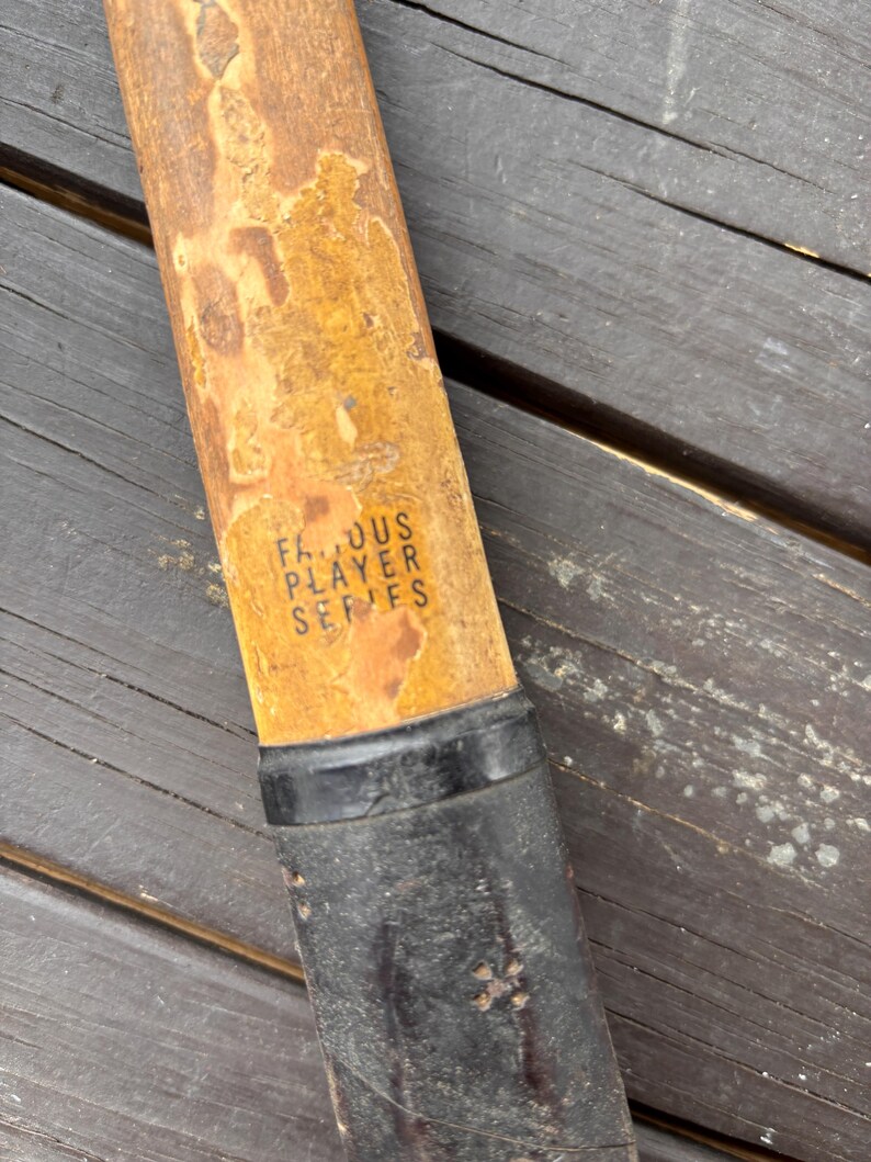 May include: Close-up of a wooden tennis racket handle with a black grip. The handle is labeled "Famous Player Series".