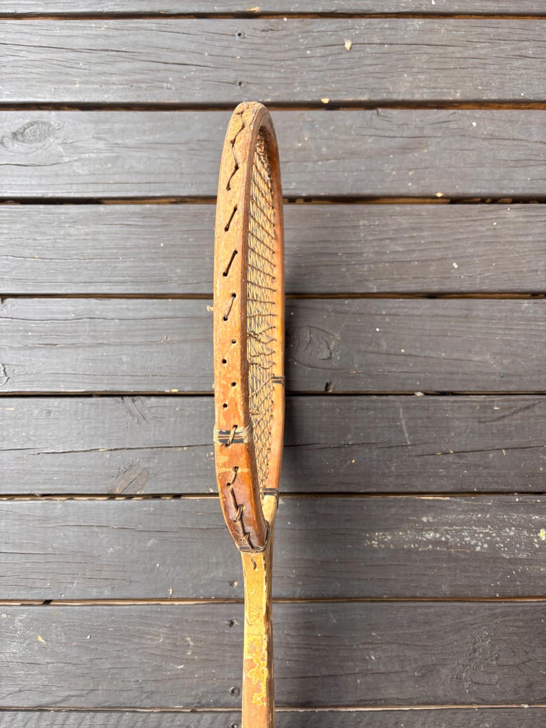May include: A vintage wooden tennis racket with a brown leather grip and a natural wood frame. The racket has a worn, distressed look, suggesting it has been used for many years.