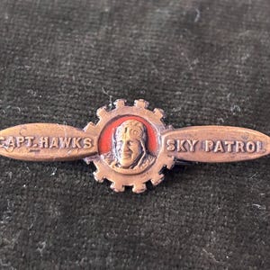 May include: A copper-toned metal pin shaped like an airplane propeller with a central gear design. The center features a relief portrait of a pilot wearing a flight cap. The text "CAPT. HAWKS" and "SKY PATROL" is embossed on the propeller blades.