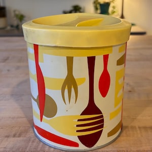 Puede incluir: Un recipiente cilíndrico amarillo y blanco con una tapa amarilla. El recipiente tiene un diseño retro con imágenes estilizadas rojas, amarillas y marrones de utensilios de cocina.