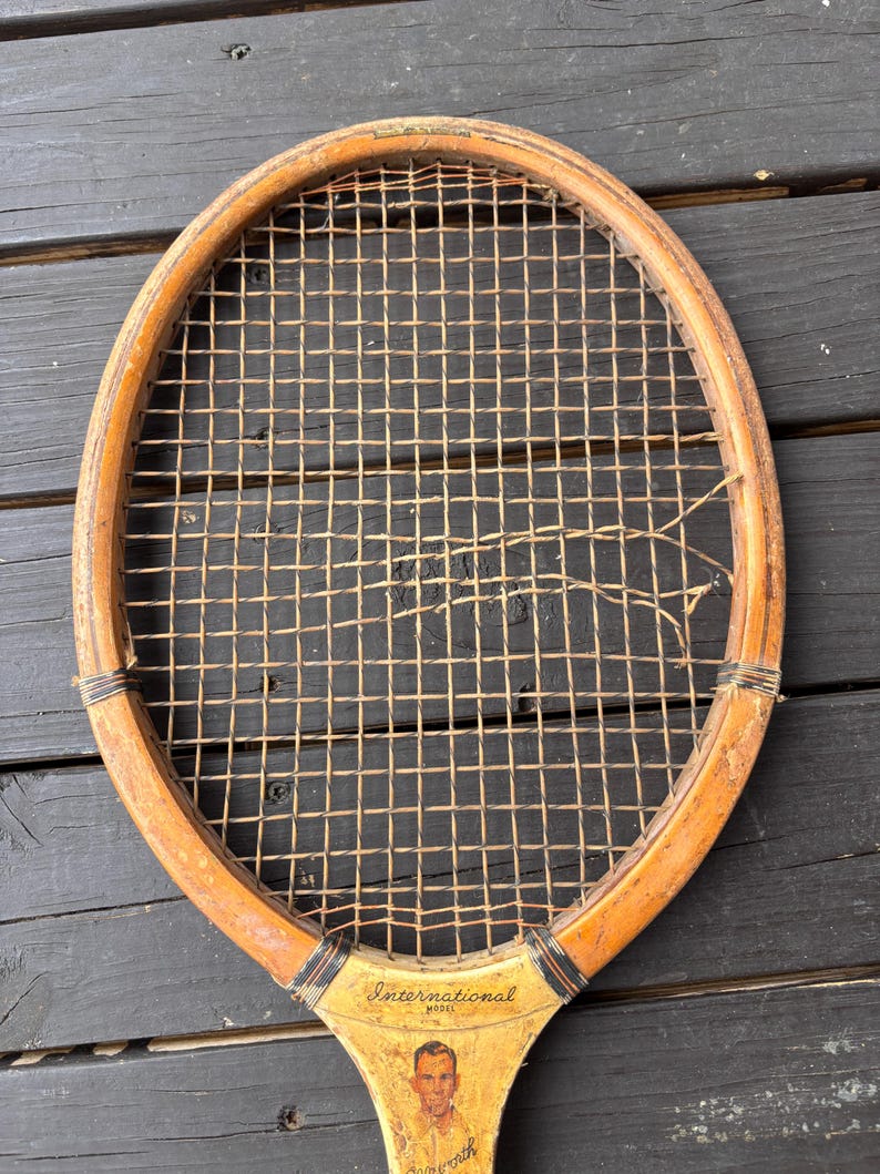 May include: A vintage wooden tennis racket with a worn stringed frame and a faded label that reads "International Model" with a picture of a man.