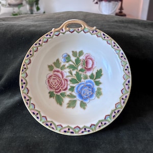 Puede incluir: Un plato redondo de cerámica blanquecina con un asa dorada. El plato presenta un diseño floral con flores rosas, azules y verdes en el centro. El borde está decorado con un borde estampado en tonos rosas, verdes y azules.