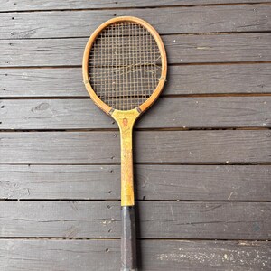 May include: A vintage wooden tennis racket with a brown wooden frame and a black handle. The racket has a worn, distressed look and a stringed net.