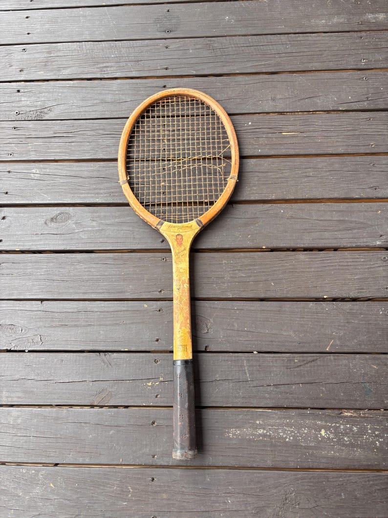 May include: A vintage wooden tennis racket with a brown wooden frame and a black handle. The racket has a worn, distressed look and a stringed net.