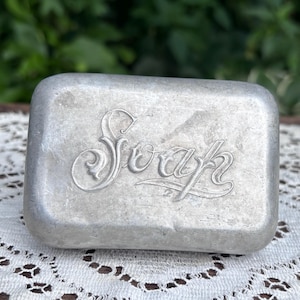 May include: A rectangular silver-toned metal soap dish or container with rounded edges. The word Soap is embossed in a decorative, cursive script across the center of the top surface. The metal has a matte, brushed finish with visible texture.