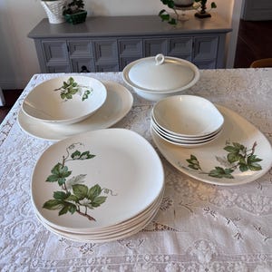 Puede incluir: Un juego de vajilla de cerámica blanca vintage con un diseño de hojas verdes y marrones. El juego incluye platos, cuencos y una fuente para servir con tapa. Los platos están apilados y los cuencos están colocados sobre un mantel blanco.