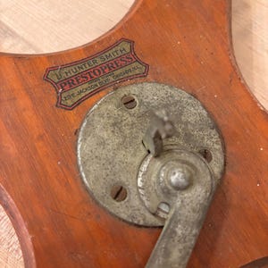 May include: A vintage wooden base with a metal lever and a label that reads "J. Hunter Smith, Patented, PrestoPress, 20 E. Jackson Blvd., Chicago, Ill."