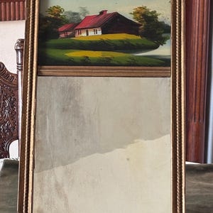 Puede incluir: Obra de arte enmarcada rectangular con una pintura de paisaje de una casa con tejado rojo, hierba verde y árboles. El marco es dorado con un diseño texturizado. La mitad inferior es un espejo.