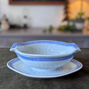 May include: White porcelain gravy boat and saucer set with blue geometric patterns. The gravy boat features a pierced design and handles. The saucer is square with rounded corners. Measurements are in centimetres.