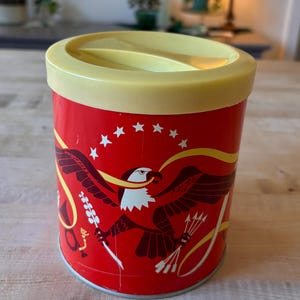 Puede incluir: Un recipiente metálico rojo vintage con tapa amarilla. El recipiente presenta un gráfico de un águila con una pancarta y estrellas. El águila está representada con una cabeza blanca y un cuerpo marrón oscuro. El recipiente es cilíndrico.