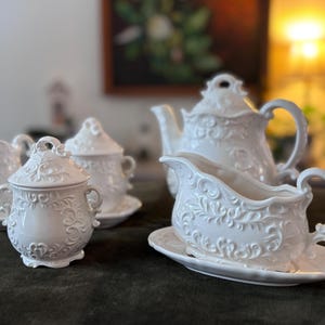 Könnte beinhalten: Ein weißes Porzellan-Teeservice mit aufwendigen floralen Details. Das Set umfasst eine Teekanne, einen Sahnekännchen, eine Zuckerdose und eine Sauciere, alle auf passenden Untertassen. Die Teile sind auf einer dunkelgrünen Oberfläche angeordnet.