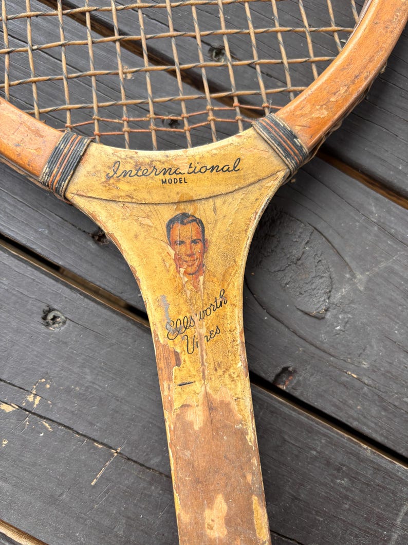 May include: Vintage wooden tennis racket with a yellowed finish. The racket has a black and white string pattern and a faded image of a man with the text "International Model Ellsworth Vines" printed on the handle.