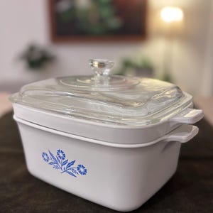 Puede incluir: Una fuente para horno blanca con tapa de cristal transparente. El plato tiene un diseño floral azul y asas blancas. La tapa tiene un pomo de cristal transparente. El plato mide aproximadamente 23 cm de largo, 23 cm de ancho y 10 cm de alto.