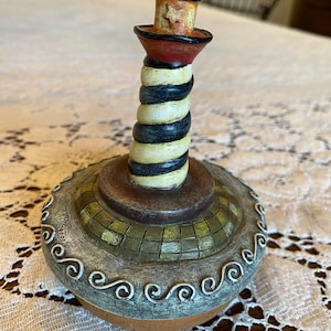 Puede incluir: Una figura decorativa de faro con una parte superior roja y negra, un cuerpo en espiral negro y amarillo y una base detallada. La base tiene un patrón de mosaico y un borde de remolinos blancos. El faro mide aproximadamente 15 cm de altura.