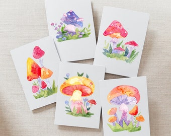 May include: Five watercolor paintings of mushrooms on white card stock. Each painting features a different mushroom with varying colors and patterns. The mushrooms are depicted with a whimsical style and are surrounded by green foliage and small flowers.