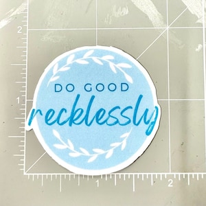 Puede incluir: Una pegatina redonda azul y blanca con un diseño de corona blanca y el texto "DO GOOD recklessly" en azul.
